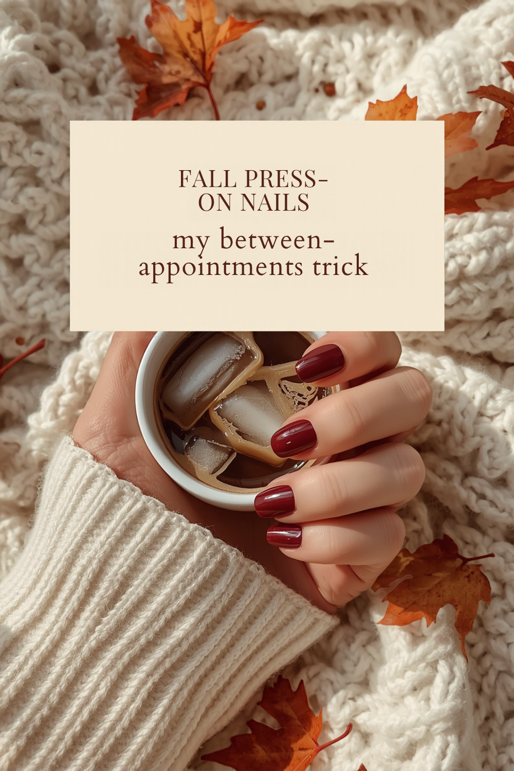 hand with short glossy wine red nails holding a cup of iced coffee on a cream cable knit blanket with orange fall leaves