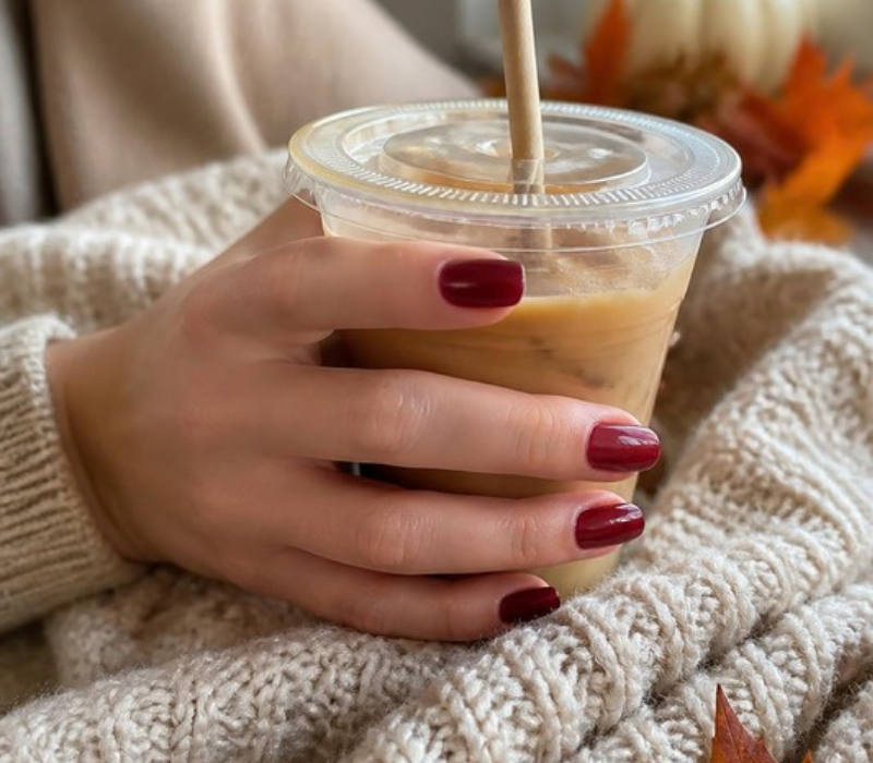 hand with short glossy deep wine red press-on nails holding a plastic cup of iced coffee over a cream knit sweater, with orange leaves and a white pumpkin behind