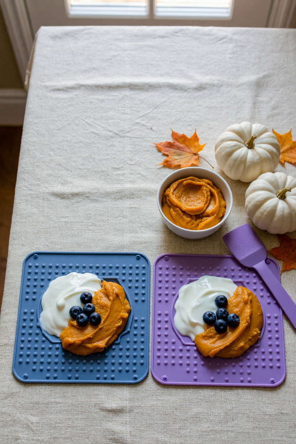 blue and purple silicone lick mats with pumpkin puree, plain yogurt and blueberries on a linen tablecloth with white pumpkins and fall leaves