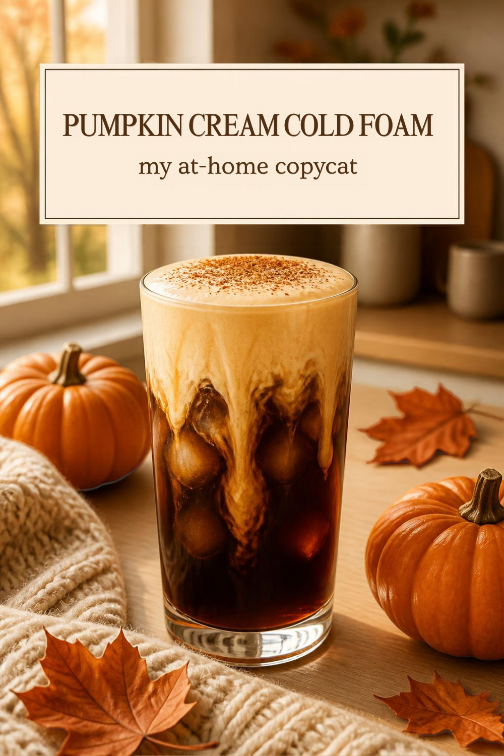 tall glass of iced cold brew topped with thick pale orange pumpkin cream cold foam dusted with spice, on a wood counter with small pumpkins, fall leaves and a cream knit blanket